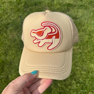 May include: Tan trucker hat featuring a red and white embroidered lion cub design. The hat has a curved brim and a mesh back. The hat is held by a hand with blue nail polish, set against a green grass background.