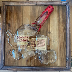 May include: A framed artwork featuring a shattered Maker's Mark bourbon bottle. The bottle's red-topped neck and fragments of glass are arranged on a wooden background, with label pieces visible. The frame is dark brown.