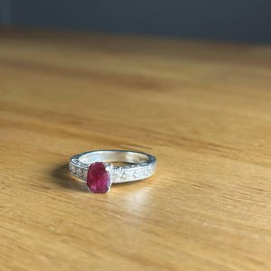 K&ouml;nnte beinhalten: Ein silberner Ring mit einem gro&szlig;en, ovalen Rubin-Edelstein. Das Ringband weist ein eingraviertes Blumenmuster auf. Der Rubin hat eine tiefrote Farbe, und der Ring liegt auf einer Holzoberfl&auml;che.