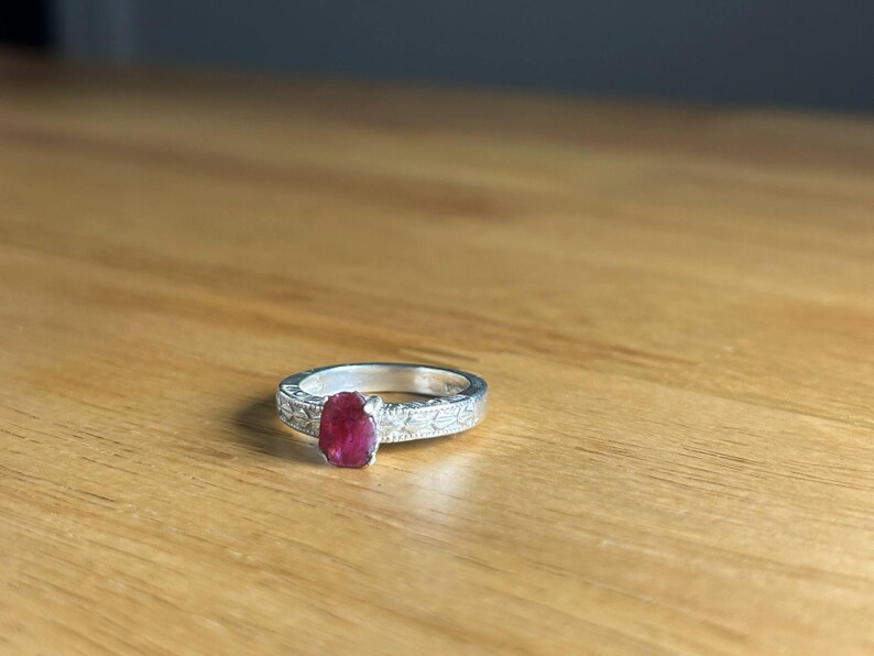 K&ouml;nnte beinhalten: Ein silberner Ring mit einem gro&szlig;en, ovalen Rubin-Edelstein. Das Ringband weist ein eingraviertes Blumenmuster auf. Der Rubin hat eine tiefrote Farbe, und der Ring liegt auf einer Holzoberfl&auml;che.