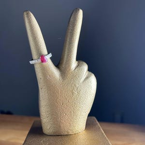K&ouml;nnte beinhalten: Eine goldfarbene Handstatue im Friedenszeichen, mit einem Rubinring am Zeigefinger. Der Ring hat ein silbernes Band mit kleinen, klaren Steinen. Die Statue steht auf einem goldfarbenen rechteckigen Sockel.