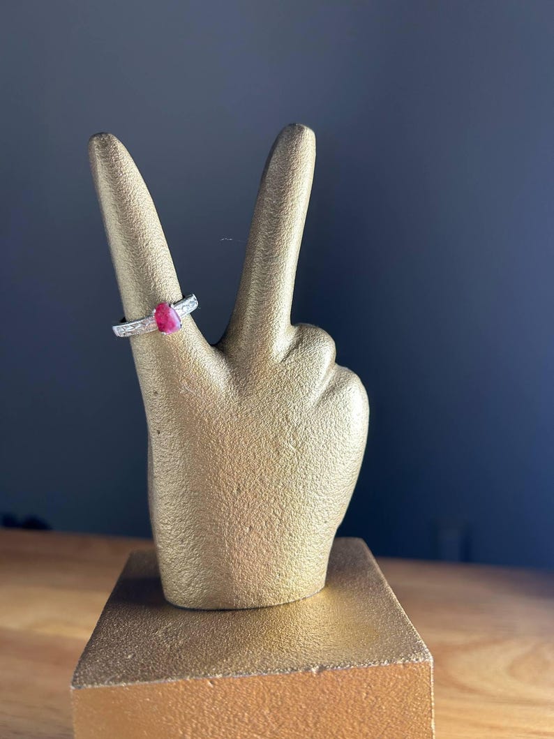 K&ouml;nnte beinhalten: Eine goldfarbene Handstatue im Friedenszeichen, mit einem Rubinring am Zeigefinger. Der Ring hat ein silbernes Band mit kleinen, klaren Steinen. Die Statue steht auf einem goldfarbenen rechteckigen Sockel.
