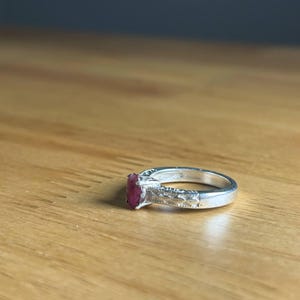 K&ouml;nnte beinhalten: Ein silberner Ring mit einem rechteckigen, tiefroten Edelstein. Das Band hat ein dekoratives Design an den Seiten. Der Ring liegt auf einer hellbraunen Holzoberfl&auml;che.