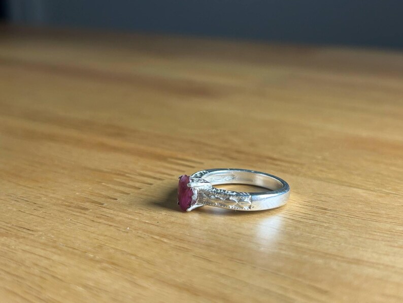 K&ouml;nnte beinhalten: Ein silberner Ring mit einem rechteckigen, tiefroten Edelstein. Das Band hat ein dekoratives Design an den Seiten. Der Ring liegt auf einer hellbraunen Holzoberfl&auml;che.