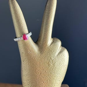 K&ouml;nnte beinhalten: Ein silberner Ring mit einem roten Edelstein wird auf einer goldfarbenen Handskulptur pr&auml;sentiert. Der Ring hat ein strukturiertes Band und einen tropfenf&ouml;rmigen Stein. Die Skulptur befindet sich vor einem dunkelblauen Hintergrund.