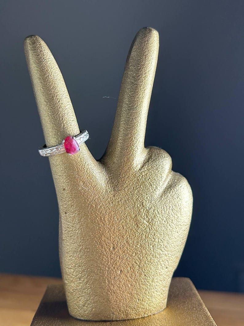 K&ouml;nnte beinhalten: Ein silberner Ring mit einem roten Edelstein wird auf einer goldfarbenen Handskulptur pr&auml;sentiert. Der Ring hat ein strukturiertes Band und einen tropfenf&ouml;rmigen Stein. Die Skulptur befindet sich vor einem dunkelblauen Hintergrund.