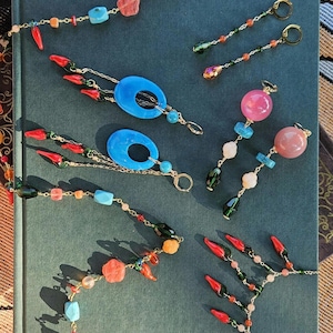 Könnte beinhalten: Eine Sammlung farbenfrohen Perlenschmucks, darunter Halsketten und Ohrringe, auf einer dunkelblauen Oberfläche. Der Schmuck besteht aus goldenen Ketten und einer Vielzahl von Edelsteinperlen in Rot-, Blau-, Rosa-, Grün- und Orangetönen.