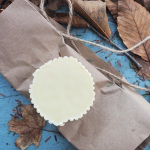 May include: A white round candle with a scalloped edge sits on a brown paper package. The package is on a blue surface with brown leaves and twigs scattered around it.