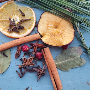 May include: A collection of dried fruits, spices, and herbs on a blue wooden surface. The ingredients include dried orange slices, dried apple slices, cinnamon sticks, star anise, cloves, bay leaves, and rosemary sprigs.
