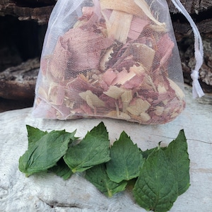 May include: A white mesh bag filled with pink and brown wood shavings, with a pile of green leaves on a light wood surface.