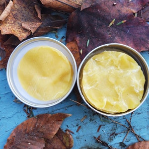 May include: Two clear glass jars with lids, filled with a yellow, waxy substance. The jars are sitting on a blue surface with brown leaves scattered around them.