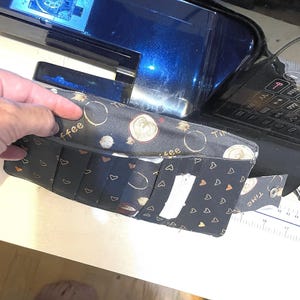 Puede incluir: Una cartera de tela negra con un estampado de temática cafetera, con diseños de tazas de café y la palabra "coffee". La cartera está abierta, revelando múltiples ranuras para tarjetas y un cierre a presión. También es visible la palabra "TIME".