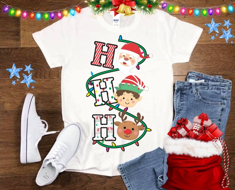 May include: White t-shirt with a red, green, and white Christmas lights design. The lights are strung around the letters "HO HO HO" and feature Santa Claus, an elf, and a reindeer.