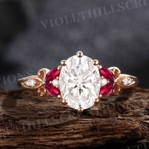 May include: Rose gold engagement ring featuring an oval-cut white center stone flanked by two red gemstones. The ring has delicate side accents with small white stones. The ring is set on a wooden surface.