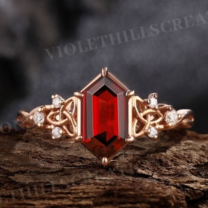 May include: A rose gold ring with a red gemstone and a Celtic knot design. The ring is set with small diamonds.