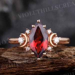 May include: Rose gold ring featuring a marquise-cut, deep red gemstone as the centerpiece. The ring is adorned with small, round white gemstones and crescent moon-shaped designs on either side of the main stone. The ring is set on a wooden surface.