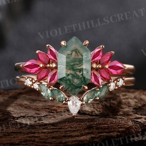 May include: A rose gold engagement ring set featuring a large, hexagonal green moss agate center stone. The ring is surrounded by vibrant pink gemstones and smaller green and white accent stones. The band is delicate and detailed, showcasing a unique design.