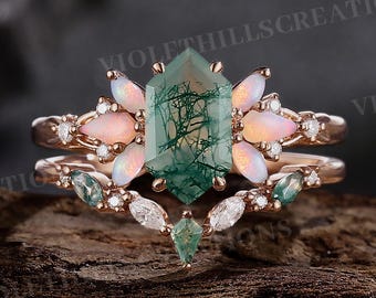 Può includere: Un set di anelli in oro rosa con una grande pietra centrale esagonale in agata muschiata verde. L'anello è affiancato da opali rosa e piccole pietre bianche. La fascia è adornata con piccoli gemme aggiuntive. Il testo "VIOLETHILLScreations" è visibile.