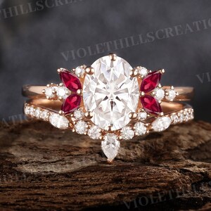 May include: A rose gold engagement ring set with an oval-cut, clear gemstone center stone, flanked by red and white accent stones. The band is encrusted with smaller white gemstones. The text "VIOLETHILLScreations" is visible.