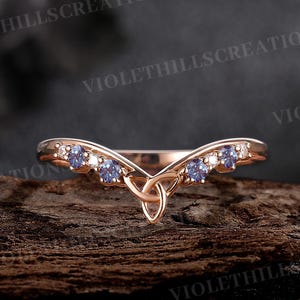 Op de afbeelding: Een delicate roségouden trouwring met een Keltisch knoopontwerp en een rij kleine blauwe edelstenen. De ring is gebogen om comfortabel om de vinger te passen.