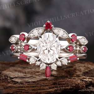 Peut inclure: Une bague de fiançailles en trois parties avec une grande pierre centrale ovale, des pierres rondes plus petites et des accents de rubis rouges.