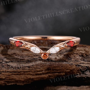May include: A rose gold ring with a curved design. The ring features red, white, and orange gemstones. The ring is set on a wooden surface. The text "VIOLETHILLScreations" is visible in the background.