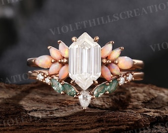 May include: A rose gold ring set featuring a central hexagonal step-cut white stone flanked by marquise-cut opals. The band includes a curved nesting piece adorned with marquise-cut green moss agate stones and small round white accent stones.