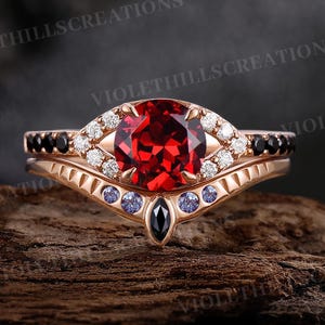 Op de afbeelding: Een roségouden ringenset met een grote, ronde, gefacetteerde rode edelsteen als middelpunt. De ring is versierd met kleine witte en zwarte edelstenen en een zwarte, druppelvormige steen. De ring ligt op een houten oppervlak.