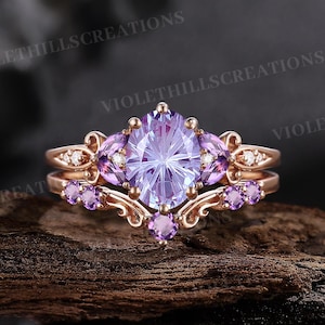 May include: A rose gold ring set featuring a large oval-cut light purple center stone flanked by two smaller round purple stones and clear accent stones. The band has an intricate scrollwork design with additional small purple stones embedded.