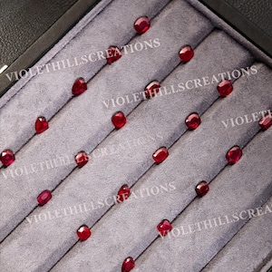 May include: A collection of small, faceted, deep red gemstones arranged in rows within a grey velvet display tray. Each stone features a cushion-cut shape with reflective surfaces. The text VIOLETHILLSCREATIONS is overlaid across the image.