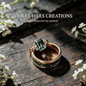 May include: A gold ring featuring a rectangular emerald-cut dark green gemstone flanked by small clear stones, resting atop a wider gold band with a textured dark green inlay. The text VIOLET HILLS CREATIONS BOLD HANDCRAFTED JEWELRY is visible.