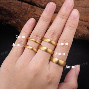 May include: A set of five gold-toned rings with a smooth, matte finish, displayed on fingers. The rings are labeled with their respective widths of 2mm, 3mm, 4mm, 5mm, and 6mm, indicating the varying thickness of each individual band.