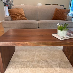 May include: A dark brown wooden coffee table with a live plant in a white pot and a book on top. The table has a rectangular top and a unique, open design with a single, wide leg on each side.