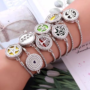 May include: A collection of silver aromatherapy bracelets with various designs, including a starfish, paw prints, a tree, and a butterfly. Each bracelet features a round pendant with a decorative design and a rhinestone border. The pendants have colored inserts.