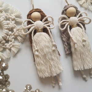 May include: Two handmade macrame angel decorations with wooden bead heads, cream-coloured bodies, and brown and grey hair. The angels have gold accents and are suspended by gold strings. Other macrame decor is visible.