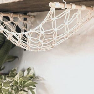 Peut inclure: Un hamac en macramé blanc suspendu à une étagère en bois. Le hamac est fait d'une corde nouée et présente un motif en losange.