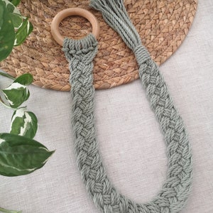 May include: A sage green macrame plant hanger with a wooden ring. The woven design creates a textured pattern. The plant hanger is designed to hold a small potted plant. The background includes a woven straw mat and green leafy plants.