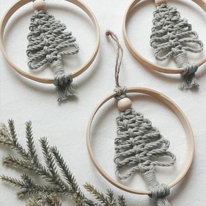 Può includere: Tre decorazioni per l'albero di Natale in macramè grigio appese ad anelli di legno. Gli ornamenti sono realizzati con una corda annodata e hanno una perla di legno in cima.