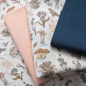 May include: A close-up of a fabric with a mushroom and bird print. The fabric is beige with brown, orange, and yellow mushrooms and birds. The fabric is folded over a pink fabric and a dark blue fabric.