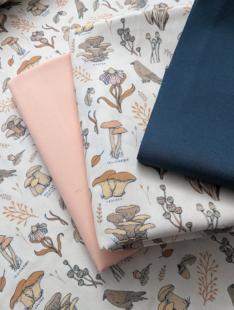 May include: A close-up of a fabric with a mushroom and bird print. The fabric is beige with brown, orange, and yellow mushrooms and birds. The fabric is folded over a pink fabric and a dark blue fabric.