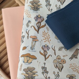 May include: A close-up of three fabric swatches. The top swatch is a white fabric with a mushroom and flower pattern. The text "HEDGEHOG", "OYSTER", "YELLOWFOOT", and "BOLETUS EDULIS" is printed on the fabric. The middle swatch is a light peach fabric. The bottom swatch is a dark blue fabric.