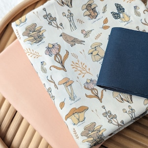 May include: Two pieces of fabric, one with a peach background and the other with a white background and a mushroom print. The mushroom print fabric has the names of the mushrooms written on it, including "Chanterelles", "Oyster", "Morels", "Yellowfoot", and "Hedgehog".