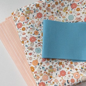 Op de afbeelding: Een stapel van drie stofvierkanten. Het bovenste vierkant is wit met een klein bloemenpatroon in tinten oranje, roze en blauw. Het middelste vierkant is effen lichtblauw. Het onderste vierkant is roze met dunne witte strepen.