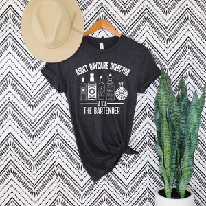Pode incluir: Camiseta cinza escuro com o texto "ADULT DAYCARE DIRECTOR A.K.A. THE BARTENDER" e ilustrações de garrafas de licor. Um chapéu bege está pendurado acima da camisa. Uma planta de cobra em vaso está à direita.