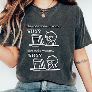 Puede incluir: Camiseta gris oscuro con texto y gráficos blancos. El texto dice "the code doesn't work... WHY?" y "the code works... WHY?" con una caricatura de una persona y una computadora.