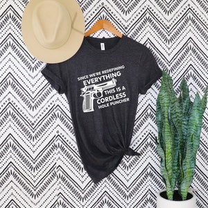 May include: Dark gray t-shirt with the text "SINCE WE'RE REDEFINING EVERYTHING THIS IS A CORDLESS HOLE PUNCHER" and a graphic of a handgun. A beige hat hangs above the shirt. The shirt is on a wooden hanger against a black and white patterned background.