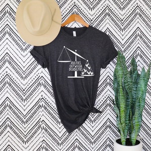 May include: Dark gray t-shirt with the text "ABILITIES OUTWEIGH DISABILITIES" in white, along with a scale graphic. A beige hat hangs above the shirt. The shirt is displayed on a wooden hanger.