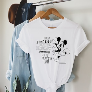 Op de afbeelding: Wit T-shirt met een zwart-witte Mickey Mouse afbeelding en de tekst "There's a great BIG BEAUTIFUL TOMORROW shining at the end of EVERY DAY!". Het T-shirt hangt aan een houten hanger.