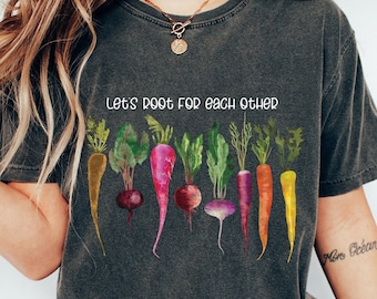 May include: Dark gray t-shirt featuring a watercolor illustration of various root vegetables, including carrots and beets. The text "Let's root for each other" is printed above the vegetables. The shirt has a vintage, washed look.