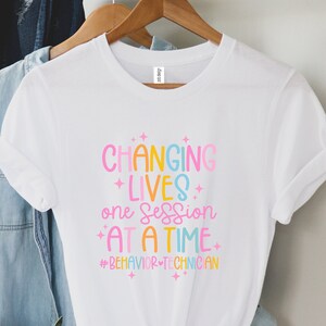 May include: White t-shirt with the text "CHANGING LIVES one session AT A TIME" in colourful letters. The shirt also includes the hashtag #BEHAVIORTECHNICIAN. The t-shirt is hanging on a wooden hanger.